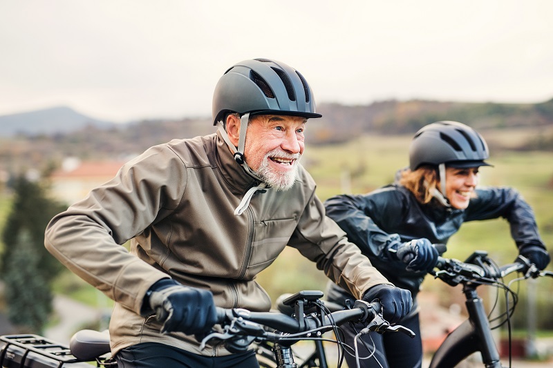 An active senior couple with helmets and electrobikes cycling outdoors on a road in nature.