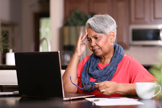 African descent, senior adult woman at home sitting at kitchen table paying bills or online banking using laptop computer. She is frustrated at the overdue charges. Paperwork and coffee cup on table.